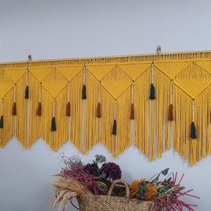 May include: A yellow macrame wall hanging with black and brown tassels. The hanging is made of a series of diamond shapes and is approximately 70 inches wide.