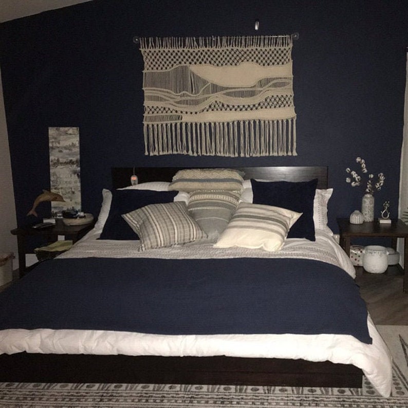 K&ouml;nnte beinhalten: Ein dunkelblaues Schlafzimmer mit einem gro&szlig;en Bett mit wei&szlig;en Bettw&auml;sche und einem dunklen Holzkopfteil. Das Bett hat mehrere Kissen in verschiedenen Mustern und Farben. Ein gro&szlig;es Makramee-Wandbild mit einem wei&szlig;en Wal-Design h&auml;ngt &uuml;ber dem Kopfteil. An beiden Seiten des Bettes stehen zwei Nachttische mit Lampen.