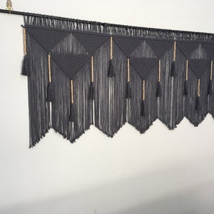 May include: Gray macrame wall hanging with wooden beads and tassels. The hanging features a geometric pattern of triangles.