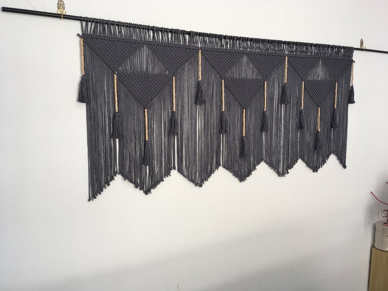 May include: Gray macrame wall hanging with wooden beads and tassels. The hanging features a geometric pattern of triangles.