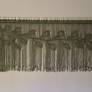 May include: A macrame wall hanging with a leafy design in a muted green color. The hanging is made of knotted cords and has a fringe at the bottom.