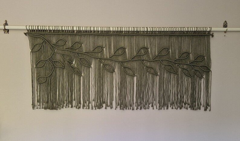 May include: A macrame wall hanging with a leafy design in a muted green color. The hanging is made of knotted cords and has a fringe at the bottom.