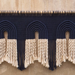 May include: A wall hanging with three black and cream macrame arches. The arches are made of thick rope and have a fringe at the bottom.
