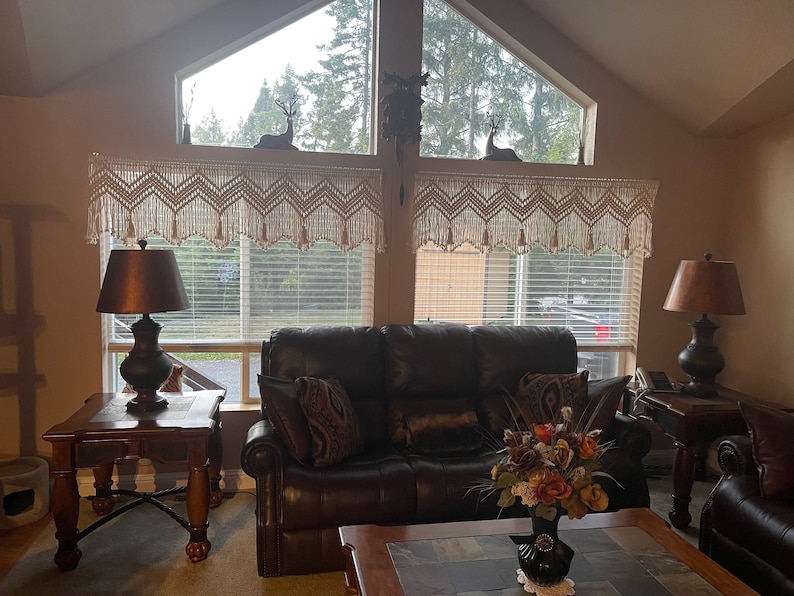 K&ouml;nnte beinhalten: Ein Wohnzimmer mit einem braunen Ledersofa, zwei Beistelltischen mit Lampen und einem Couchtisch mit einer Blumenvase. Die Fenster haben wei&szlig;e H&auml;kelvorh&auml;nge und Jalousien.