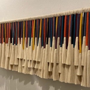 Puede incluir: Un tapiz de macram&eacute; de pared colorido con una variedad de hilos de lana de diferentes colores y borlas blancas.