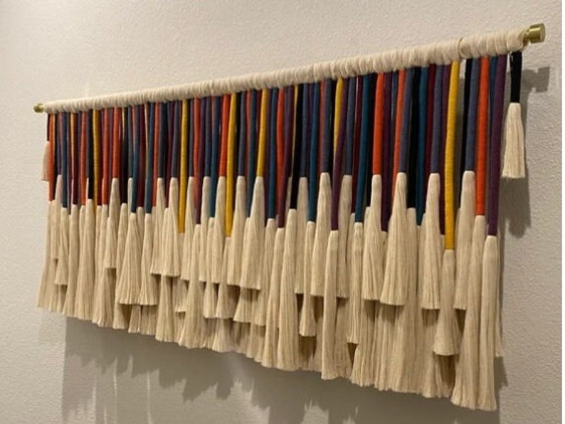 Puede incluir: Un tapiz de macram&eacute; de pared colorido con una variedad de hilos de lana de diferentes colores y borlas blancas.