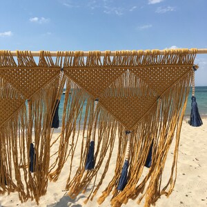 May include: Three macrame wall hangings with a geometric pattern in a mustard yellow colour with blue tassels hanging from the bottom. The hangings are displayed against a backdrop of a sandy beach and blue ocean.