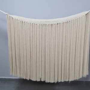 May include: A white macrame wall hanging with long, thin strands of yarn. The hanging is in the shape of a curved rectangle.