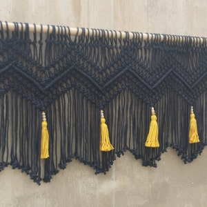May include: Black macrame wall hanging with a geometric pattern and yellow tassels. The hanging is attached to a wooden dowel.