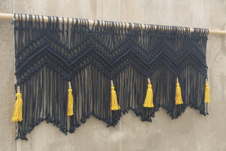 May include: Black macrame wall hanging with a geometric pattern and yellow tassels. The hanging is attached to a wooden dowel.