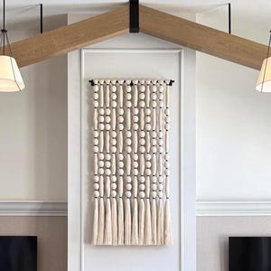 May include: A cream-colored wall hanging with a beaded design and long tassels. The wall decor features alternating rows of round and oblong beads. The piece is mounted on a white wall, and the room has a modern aesthetic.