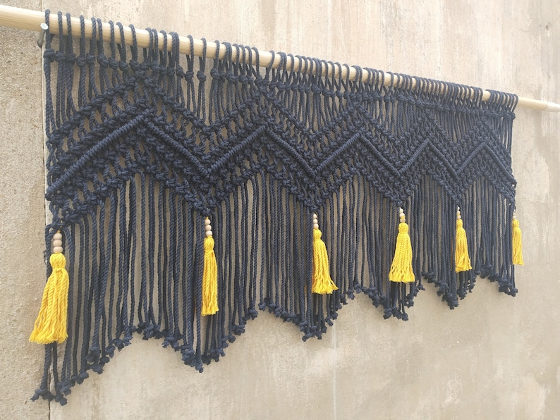 May include: Black macrame wall hanging with a geometric pattern and yellow tassels. The hanging is attached to a wooden dowel.