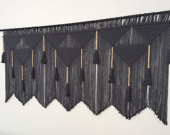 May include: A gray macrame wall hanging with wooden beads and tassels. The hanging features a geometric pattern of triangles.