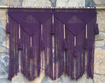 May include: Purple macrame wall hanging with wooden beads and tassels. The hanging features a geometric pattern with a triangular shape.