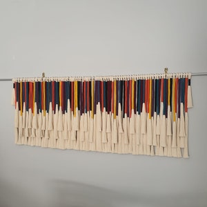 Puede incluir: Un tapiz de macram&eacute; de pared colorido con una variedad de hilos de lana de diferentes colores, incluyendo rojo, naranja, amarillo, verde, azul, morado y negro. Los hilos est&aacute;n atados juntos con borlas blancas.
