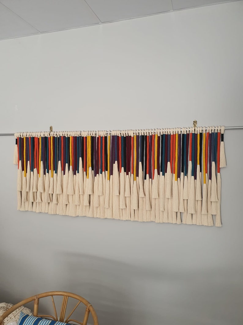 Puede incluir: Un tapiz de macram&eacute; de pared colorido con una variedad de hilos de lana de diferentes colores, incluyendo rojo, naranja, amarillo, verde, azul, morado y negro. Los hilos est&aacute;n atados juntos con borlas blancas.