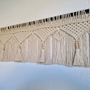 May include: Cream-colored macrame wall hanging with geometric diamond patterns and fringe. Wooden beads and tassels add detail to this handcrafted textile art piece. The macrame is mounted on a black rod.