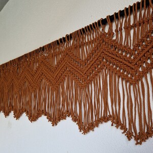 May include: A brown macrame wall hanging with a geometric pattern and fringe. The hanging is made of natural fiber rope and is attached to a black rod.