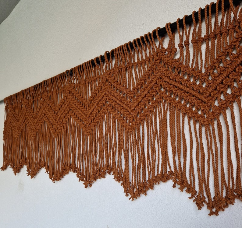 May include: A brown macrame wall hanging with a geometric pattern and fringe. The hanging is made of natural fiber rope and is attached to a black rod.