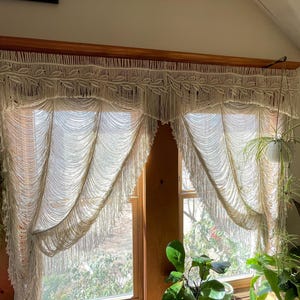 May include: Cream-colored macrame window curtains. The curtains are made of woven cotton with a fringe design. The curtains are hanging in front of a window, with sunlight shining through. The curtains are a decorative element for a home.