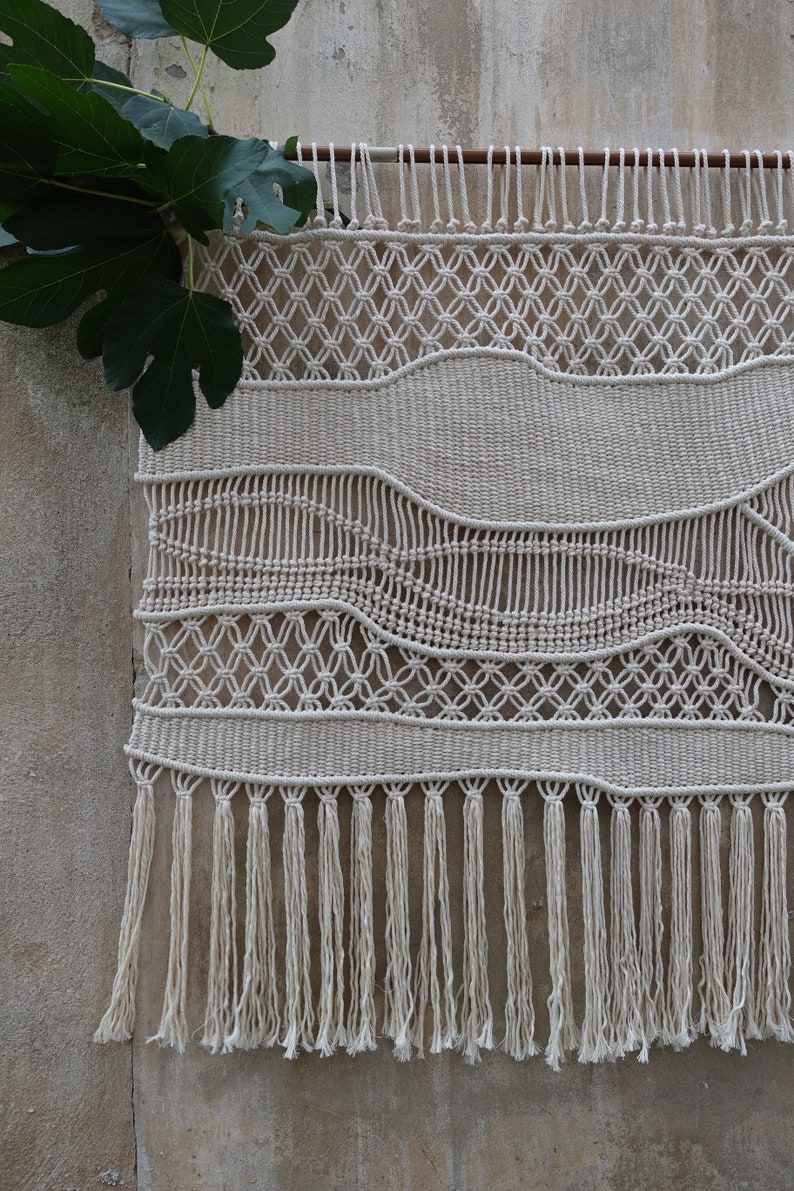 K&ouml;nnte beinhalten: Ein wei&szlig;er Makramee-Wandbehang mit geometrischem Muster und Fransen. Der Behang ist aus nat&uuml;rlichem Baumwollgarn gefertigt und ein beliebtes Dekoelement im Boho-Stil.