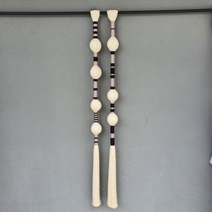 Puede incluir: Dos colgantes de pared verticales decorativos. Cada uno presenta una serie de esferas y borlas blanquecinas, alternando con rayas negras y gris claro. Los colgantes están suspendidos de una barra negra contra una pared gris.