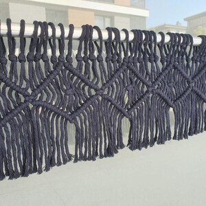 May include: A dark charcoal gray macrame wall hanging featuring a repeating diamond pattern created with thick cotton cord. The piece is suspended from a silver metal rod and finished with long, straight fringe along the bottom edge.