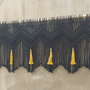 May include: Black macrame wall hanging with a geometric pattern and yellow tassels. The hanging is attached to a wooden dowel rod.