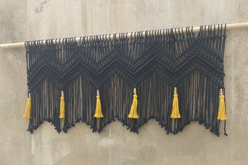 May include: Black macrame wall hanging with a geometric pattern and yellow tassels. The hanging is attached to a wooden dowel rod.