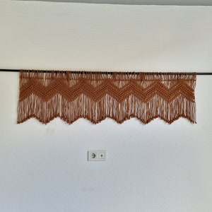 May include: A brown macrame wall hanging with a chevron pattern and fringe. The hanging is attached to a black rod with gold brackets.