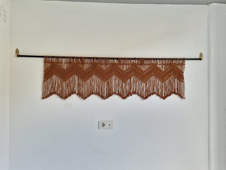 May include: A brown macrame wall hanging with a chevron pattern and fringe. The hanging is attached to a black rod with gold brackets.