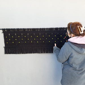 May include: A black macrame wall hanging with a geometric pattern and fringe. The macrame is decorated with small yellow beads. The wall hanging is displayed on a black rod against a grey wall.