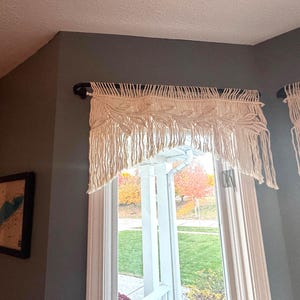 May include: Cream-colored macrame window valance with a geometric design, hanging from a black rod. The valance has a fringed bottom and is installed above a window with a view of trees.