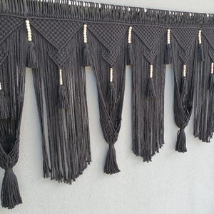 May include: Black macrame wall hanging with a geometric design. The handmade piece features a series of diamond-shaped patterns, tassels, and wooden beads. The wall decor is a stylish addition to any room, adding texture and a bohemian aesthetic.