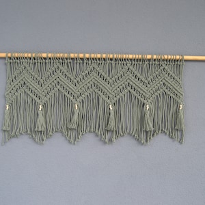 以下が含まれることがあります： 木製の棒と金色のビーズが付いた緑色のマクラメの壁掛け。壁掛けは、タッセルが付いた幾何学模様をしています。
