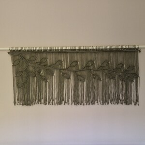 May include: A macrame wall hanging with a leafy design in a muted green color. The hanging is made with thick cord and has a fringe at the bottom.