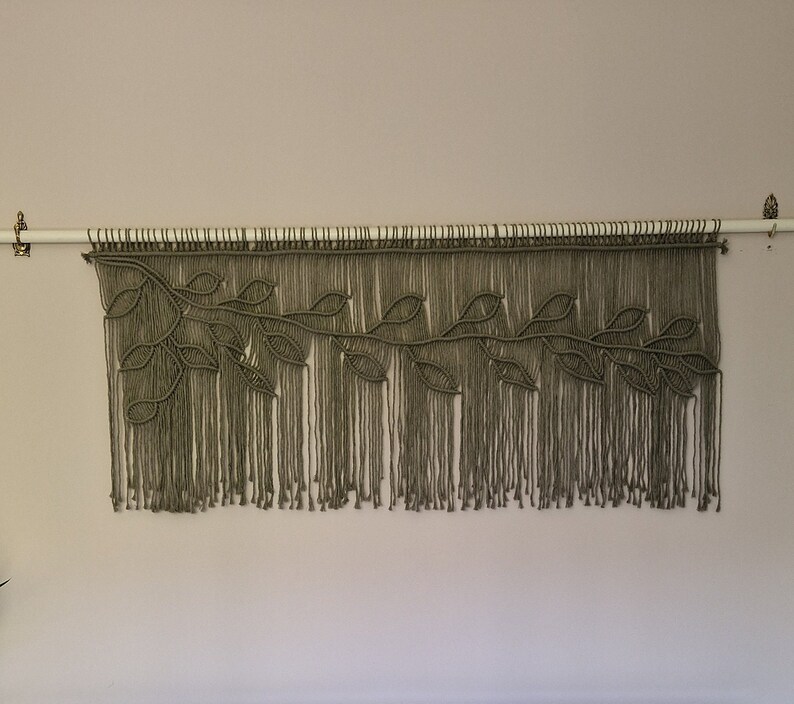 May include: A macrame wall hanging with a leafy design in a muted green color. The hanging is made with thick cord and has a fringe at the bottom.