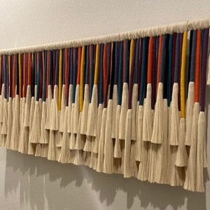 Puede incluir: Un tapiz de pared hecho de hilo blanco con hebras de colores tejidas. Las hebras son en tonos de rojo, naranja, amarillo, azul y morado. El hilo est&aacute; atado en borlas en la parte inferior.
