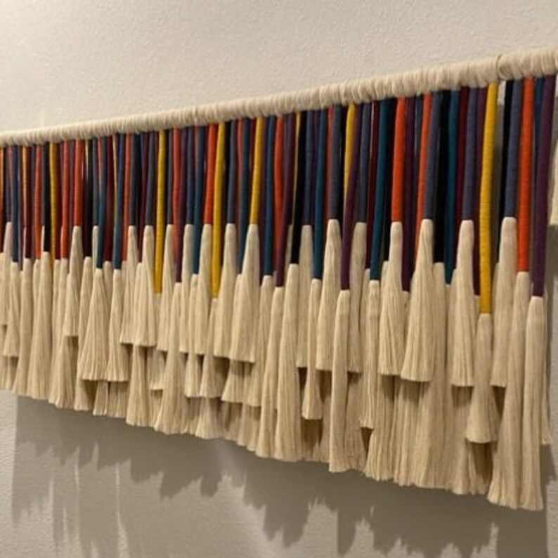 Puede incluir: Un tapiz de pared hecho de hilo blanco con hebras de colores tejidas. Las hebras son en tonos de rojo, naranja, amarillo, azul y morado. El hilo est&aacute; atado en borlas en la parte inferior.