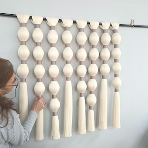 May include: A wall hanging consisting of eight vertical strands of off-white yarn, each featuring a series of rounded, bulbous sections separated by smooth, light brown cylindrical bands, all suspended from a horizontal black metal rod.