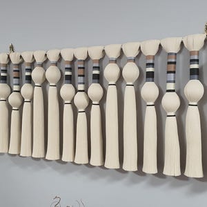 May include: A decorative wall hanging featuring multiple cream-colored tassels suspended from a silver rod. Each tassel has a cylindrical body with a decorative band of black, gray, and brown stripes. The wall hanging is mounted on a gray wall.