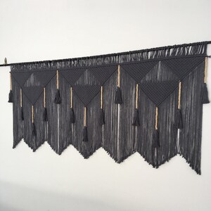 May include: A gray macrame wall hanging with a geometric pattern and wooden beads. The hanging has a fringe of tassels at the bottom.