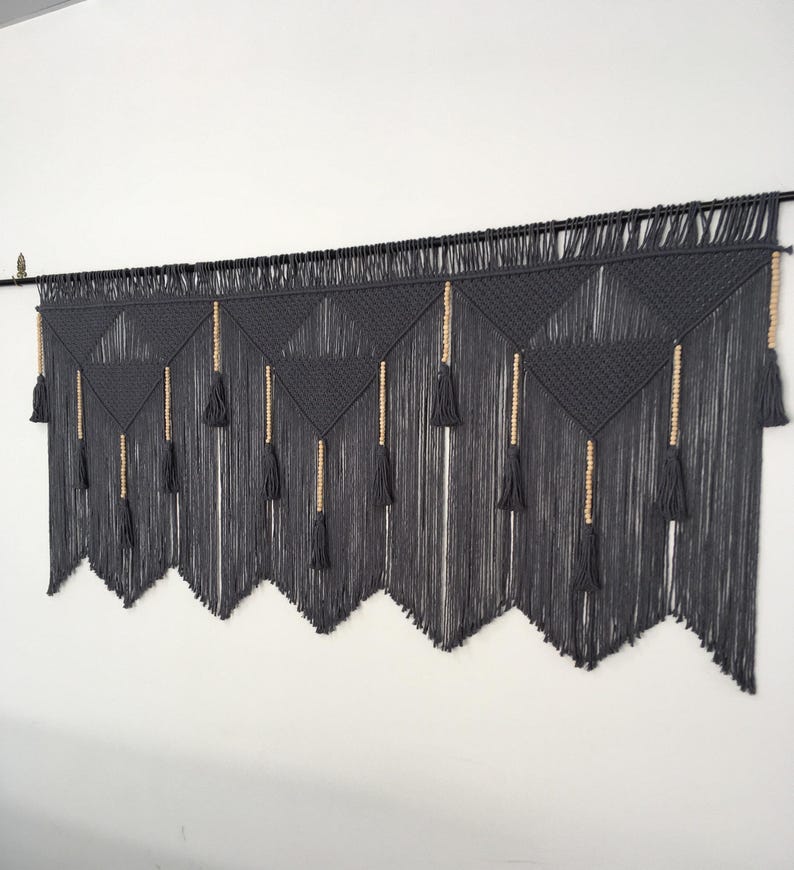 May include: A gray macrame wall hanging with a geometric pattern and wooden beads. The hanging has a fringe of tassels at the bottom.