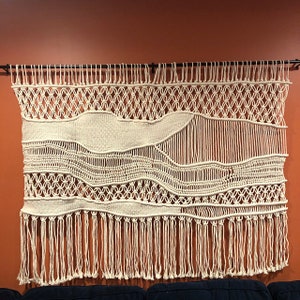 K&ouml;nnte beinhalten: Ein gro&szlig;er, wei&szlig;er Makramee-Wandbehang mit geometrischem Muster und Fransen. Der Wandbehang ist aus Naturfasern gefertigt und hat einen Boho-Stil.