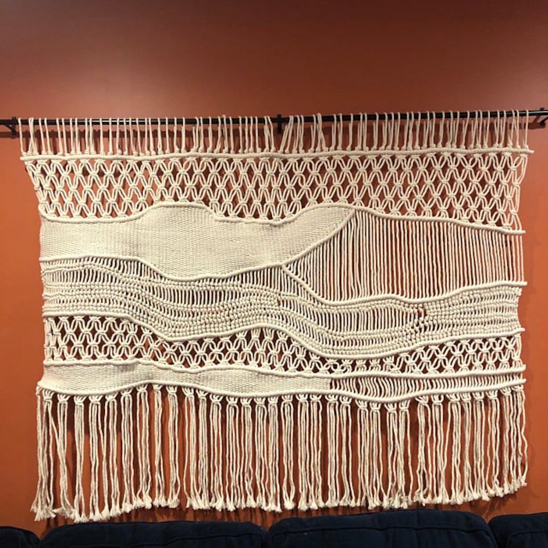 K&ouml;nnte beinhalten: Ein gro&szlig;er, wei&szlig;er Makramee-Wandbehang mit geometrischem Muster und Fransen. Der Wandbehang ist aus Naturfasern gefertigt und hat einen Boho-Stil.