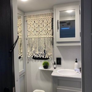 Könnte beinhalten: Eine weiße Toilette mit geschlossenem Deckel in einem kleinen Badezimmer mit weißen Wänden und einem weißen Waschtisch mit Waschbecken. Auf der Ablage steht eine kleine Pflanze und über dem Waschbecken hängt ein Wandbehang aus Makramee.
