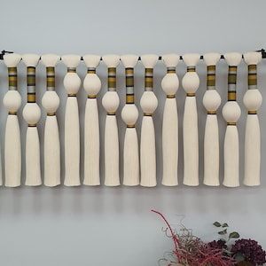 Könnte beinhalten: Ein Wandbehang aus weißen Quasten mit goldenen und schwarzen Streifenbändern. Die Quasten hängen an einer schwarzen Stange.