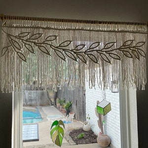 Pode incluir: Uma cortina de macramé branca com um design de folhas pendura em uma janela. A janela dá para um quintal com uma piscina, plantas e uma parede de tijolos brancos. Duas peças de vitral penduram na janela, uma com a forma de uma folha e a outra com a forma de um cubo.