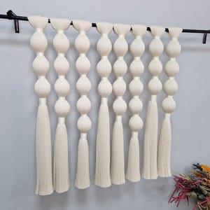 May include: A wall hanging featuring eight vertical strands of off-white fiber, each composed of a series of stacked, rounded, bulbous shapes that transition into long, straight, fringed tassels at the bottom, all suspended from a black rod.