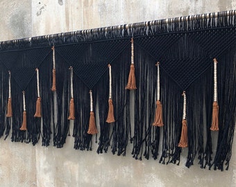 May include: A large black macrame wall hanging suspended from a wooden rod. The design features intricate knotted geometric triangle patterns, long black fringe, and vertical accents of light wooden beads paired with brown tassels throughout.